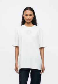 Bílé bavlněné oversize tričko s krátkými rukávy, na prsou se nachází kulatý logotyp. Hladká textura a volný střih.