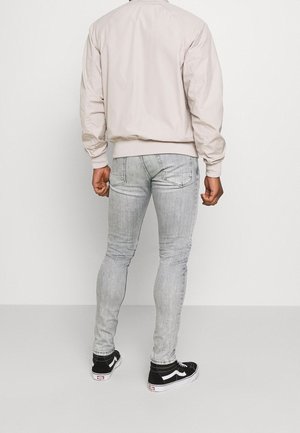 Homme portant une veste beige clair, un jean slim gris clair et des baskets montantes noires, debout face à un mur blanc uni.