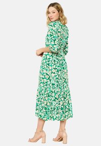 Abito midi floreale verde con maniche lunghe, vita stretta e gonna a più strati. Caratteristiche notevoli di motivi floreali beige e tessuto leggero e fluido.