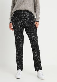 Sorte bukser med mønster og løs pasform, præget af et tekstureret, abstrakt design, parret med hvide sneakers og en lysegrå sweater.