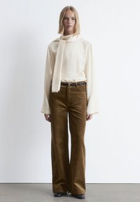 Cream blouse with long sleeves and scarf detail, paired with brown corduroy flared pants. Model stands against a plain background.