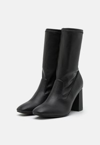 Botas de tobillo de cuero negro con un alto tacón cuadrado, punta afilada y paneles laterales elásticos para facilitar su uso. Textura suave y diseño minimalista.