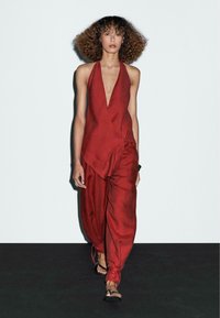 Red linen jumpsuit with deep V-neck and wrap design; loose-fitting trousers; model wears flat sandals. White background, black floor.