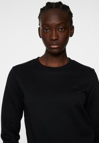 Svart sweatshirt med rundad hals och subtil logotypdetalj. Har en slät textur och en figurnära silhuett, lämplig för avslappnad klädsel.