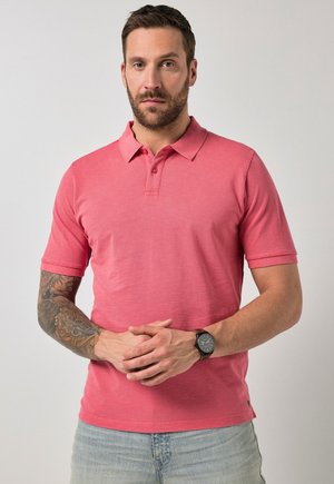 Mężczyzna z brodą i wytatuowaną prawą ręką, ubrany w różową koszulkę polo i jasnoniebieskie dżinsy, stojący z złączonymi dłońmi na wysokości pasa.