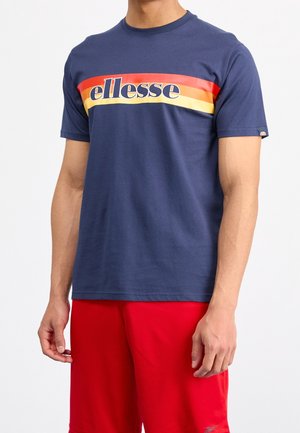 Homme portant un T-shirt Ellesse bleu marine avec des rayures rouge, orange et jaune, ainsi qu'un short de sport rouge, sur un fond blanc uni.