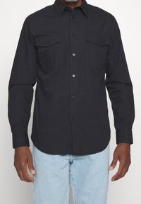 Camisa negra de manga larga con botones en la parte delantera, dos bolsillos en el pecho y un dobladillo curvado. Fabricada con tela de tejido y con una textura suave. Diseño clásico.