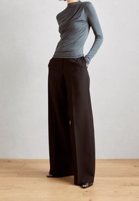 Person i en figurnära långärmad grå topp och högmidjade vida svarta byxor, stående på trägolv mot en enfärgad vägg.