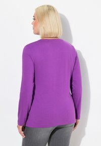 Lila Langarmshirt, von hinten gezeigt, mit Rundhalsausschnitt und glattem, leichtem Stoff. Kombiniert mit grauen Hosen.