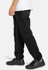 Pantalones de mezclilla negros con costuras visibles, ajuste relajado y detalles en las costuras laterales, combinados con zapatillas deportivas negras.