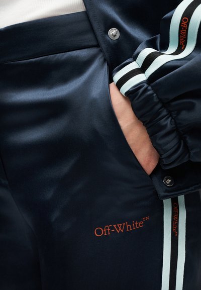 Χέρι στην τσέπη με ναυτικομπλε σατέν παντελόνι φόρμας Off-White™ και ταιριαστό μπουφάν με ανοιχτό μπλε, μαύρες ρίγες και πορτοκαλί κείμενο με λογότυπο.