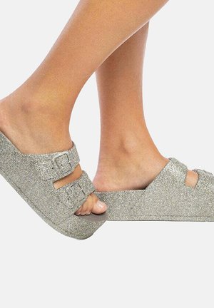 Gros plan de deux pieds portant des sandales argentées scintillantes avec des boucles, mettant en valeur le design et la texture de la chaussure sur un fond uni.