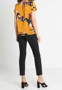 Žlutá květinová blůza s krátkými rukávy a klíčovou dírkou na zádech; spárovaná s černými přiléhavými džíny a černými sandály na blokovém podpatku.