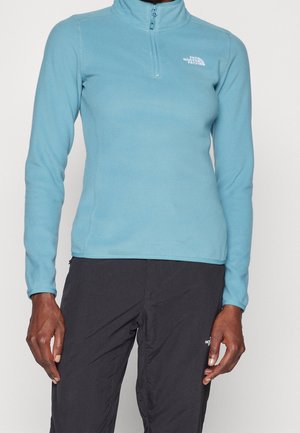 Osoba ubrana w jasnoniebieski polar z suwakiem do ćwiartki i czarne spodnie, oba z logo The North Face, stojąca na jednolitym tle.