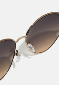 Cat-eye sunglasses with a gold metal frame, gradient brown lenses, and clear nose pads. Sleek design complements oversized shape.