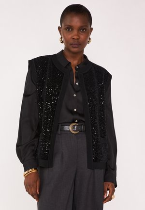 Gilet senza maniche nero con paillettes frontali, scollatura rotonda, abbinato a una camicia nera con bottoni e pantaloni grigi a vita alta. Visibili dettagli in gioielli dorati.