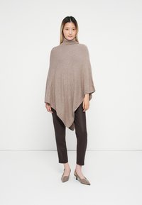 Poncho bege tricotado com gola alta, apresentando um design fluido e uma bainha pontuda, combinado com calças castanhas e sapatos claros.