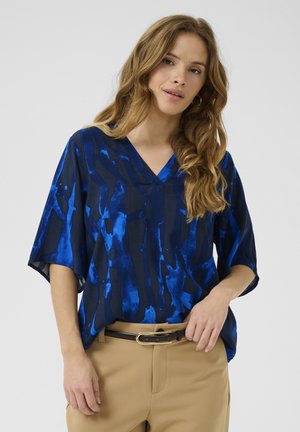 Blouse bleu à col en V avec des motifs abstraits bleu marine foncé et bleu clair, coupe décontractée, manches courtes, portée sur un pantalon beige avec une ceinture noire.