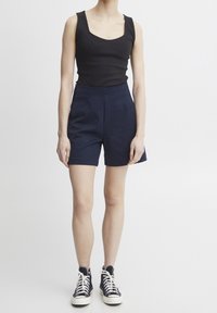 Zwarte mouwloze top met vierkante halslijn, gecombineerd met marineblauwe high-waisted shorts. Model draagt zwarte hoge sneakers met witte zolen.