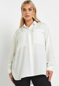 Camicia bianca a maniche lunghe con bottoni, due tasche sul petto, vestibilità rilassata e tessuto leggero e testurizzato. Presenta un colletto.