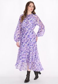 Lungo vestito floreale viola con tessuto trasparente, maniche a sbuffo, orlo arricciato e scollatura con cordoncino. Indossato con stivaletti neri alla caviglia.