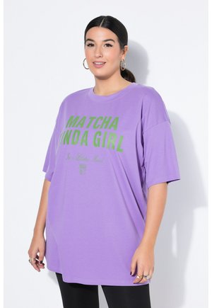 Kobieta z ciemnymi włosami związanymi w kucyk, nosząca złote kolczyki koła i luźny fioletowy T-shirt z zielonym napisem "MATCHA KINDA GIRL."