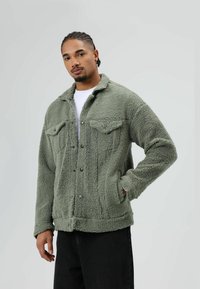 Green, fluffy fleece jacket with a collared neck, button closure, two chest pockets, and a textured surface. Worn with a white shirt.