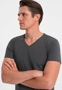Tmavě šedé tričko s výstřihem do V vyrobené z měkké tkaniny, s krátkými rukávy a hladkým povrchem, nošené mužským modelem se založenýma rukama.