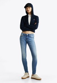 Sudadera corta de punto marinero, camiseta blanca debajo, jeans slim-fit de color azul claro y zapatillas beige con suelas marrones. Gorra negra en la cabeza.