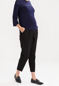 Mujer vistiendo un suéter azul marino con encaje, pantalones negros cortos y mocasines negros de punta, de pie con las manos en los bolsillos.