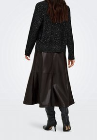 Femme aux longs cheveux bruns portant un pull noir scintillant, une jupe midi en cuir brun foncé et des bottes à talons noires, vue de dos.