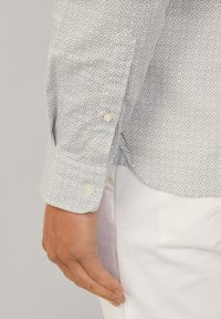 Grau gemustertes Button-Up-Hemd mit langen Ärmeln, das ein Rautenmuster zeigt, und einem weißen Knopf an den Manschetten. Getragen mit weißen Hosen.