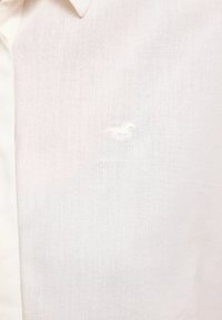 Chemise blanche à col avec un logo brodé subtil représentant un cheval et un cavalier sur la poitrine, texture du tissu visible.