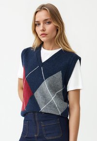 Maglione senza maniche blu navy con motivo a losanghe in rosso, grigio e bianco. Design a V, lavorazione a maglia testurizzata, indossato sopra una camicia bianca.