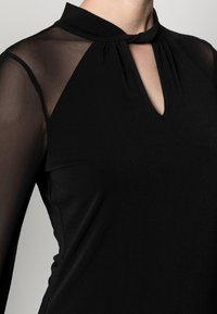 Abito nero con collo alto e maniche trasparenti. Presenta un'apertura a forma di chiave all'altezza del collo e un corpo liscio e strutturato.