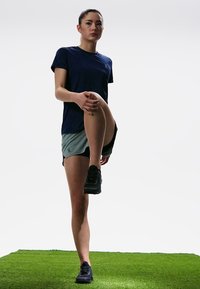 T-shirt de sport bleu marine associé à un short gris clair, mannequin effectuant un étirement des jambes sur du gazon, portant des chaussures de course noires.