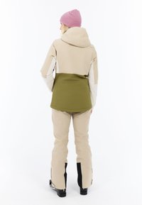 Lichtbeige en olijfgroene sneeuwjack met een capuchon, gecombineerd met bijpassende beige sneeuwbroek. Beschikt over contrasterende witte mouwen en zwarte accenten.