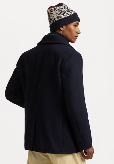 Manteau en laine bleu marine avec un large col, des plis au dos, et un bonnet en tricot texturé avec un motif rouge, blanc et noir.
