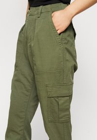 Persoon in olijfgroene cargobroek met zakken, hand gedeeltelijk in de voortas, gecombineerd met een zwart topje tegen een eenvoudige achtergrond.