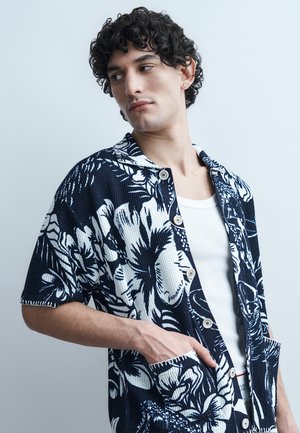 Jeune homme aux cheveux bouclés portant une chemise à manches courtes noire et blanche à motifs floraux par-dessus un débardeur blanc, regardant sur le côté.