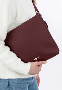 Burgundi värvi õlakott, millel on sile tekstuur, dekoratiivsed kuldsed detailid ja lai rihm. Sobitatud valge karvase kampsuni ja siniste teksadega.