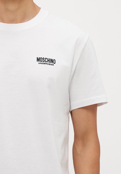 Balta medvilninė marškinėliai trumpomis rankovėmis su juodu išsiuvinėtu "Moschino Underwear" logotipu kairėje krūtinės pusėje. Lygi tekstūra.