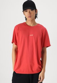Tricou roșu din bumbac cu mâneci scurte, cu un mic logo alb Levi's pe piept. Modelul poartă o șapcă neagră de baseball.
