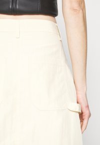 Personne portant un haut court noir et une jupe crème taille haute avec de grandes poches arrière et un détail de sangle latérale.