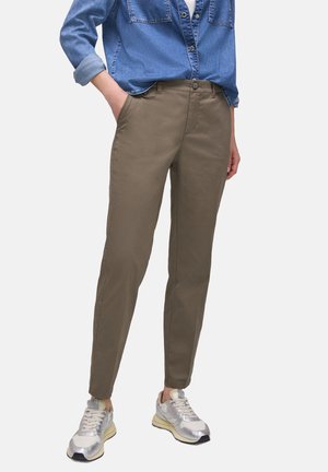 Osoba ubrana w podwiniętą jeansową koszulę, oliwkowozielone wąskie spodnie z rękami w kieszeniach oraz białe i srebrne trampki stojąca na jednolitym tle.