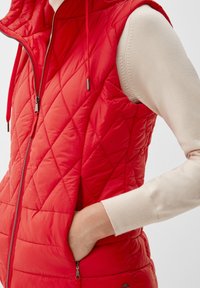 Gilet matelassé rouge avec capuche, doté d'une fermeture éclair et de poches latérales. Fabriqué en tissu léger et brillant avec une texture lisse.