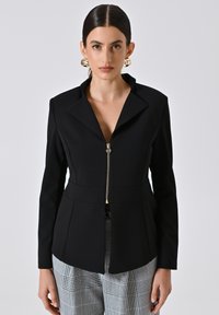 Blazer noir cintré avec un col cranté, fermeture éclair à l'avant et coupe structurée ; associé à un pantalon à carreaux gris. Texture de tissu lisse.