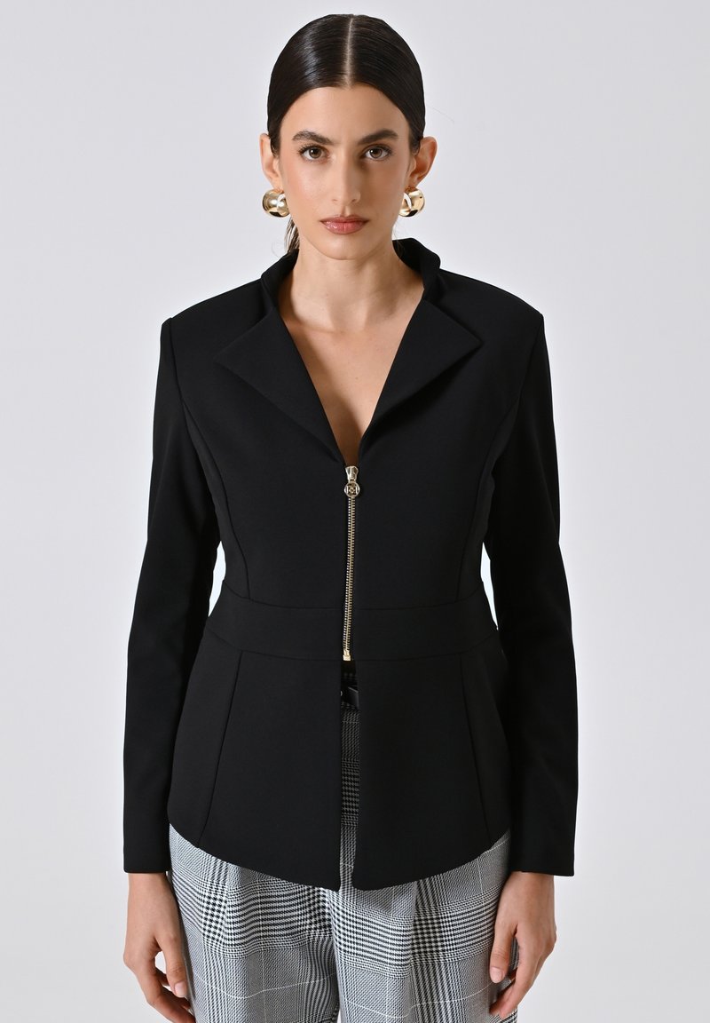 Schwarzer taillierter Blazer mit Reverskragen, vorne mit Reißverschluss und strukturiertem Schnitt; kombiniert mit grauen karierter Hose. Glatte Stofftextur.