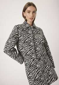 Mladá žena v černobílé zebra vzoru sako se zipem vpředu a odpovídající sukní, stojící na jednobarevném pozadí.