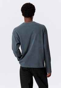 Majica s dugim rukavima u tamnoplavoj boji, izrađena od mekanog materijala, s opuštenim krojem, okruglim izrezom i spuštenim ramenima. Nema vidljivih uzoraka ili naglasaka.